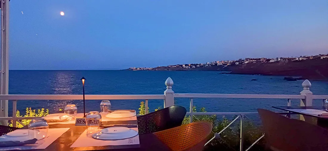 A seaside taverna terrace at dusk, table set as the full moon rises over the bay