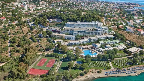 The Zeus Dolce resort from above — buildings, pool and tennis courts on a green slope above Vravrona Bay