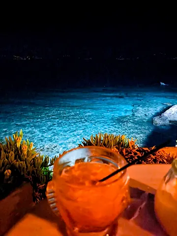 A cocktail on a seaside table at night, the lit-up Aegean just below